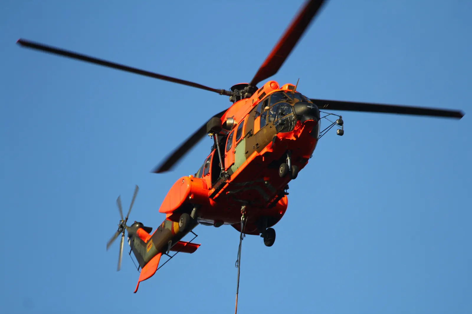 Un helicóptero de color naranja fluorescente sobrevuela una ladera boscosa cubierta de humo durante un incendio forestal.
