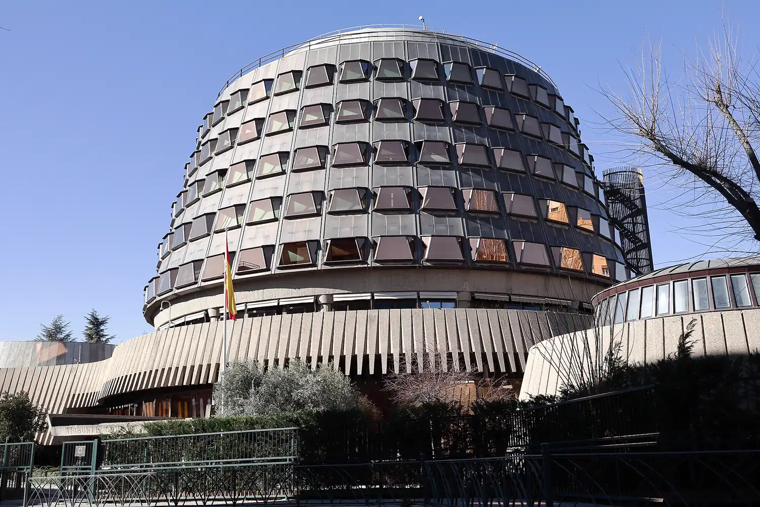 Fachada acristalada y circular de la sede del Tribunal Constitucional en Madrid, con la bandera de España ondeando ante la entrada.