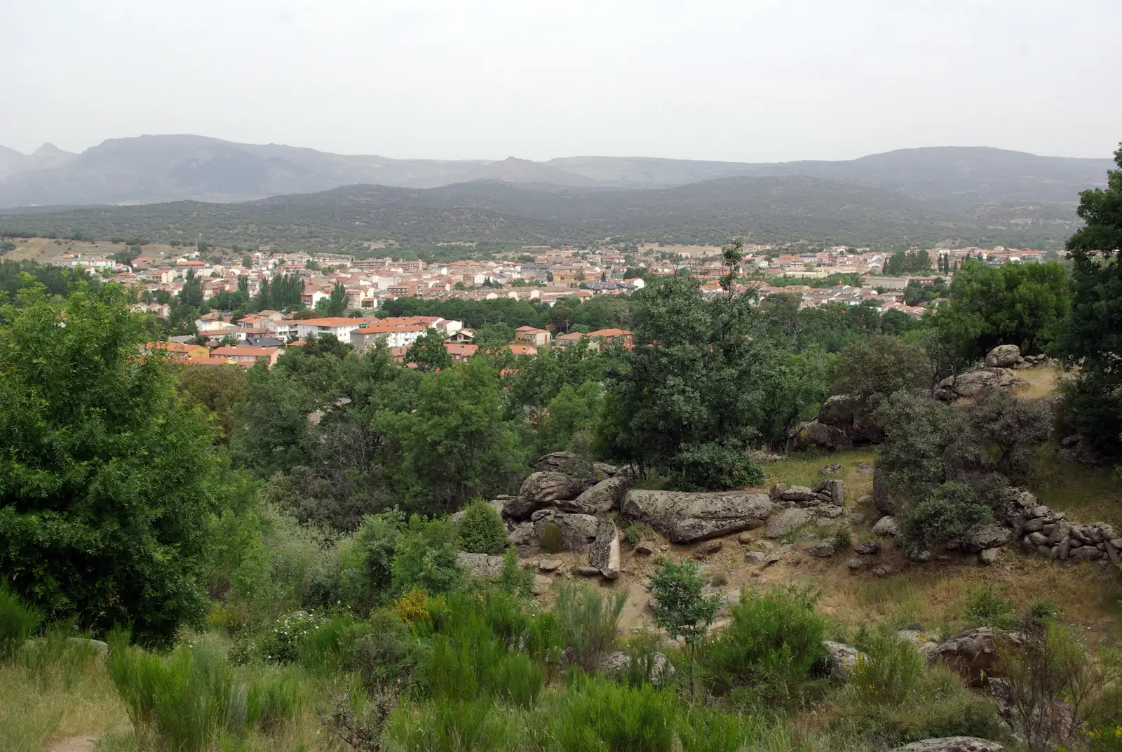 Vista de Navaluenga (Ávila) desde una ladera de encinar y roquedo, con el caserío del pueblo en el valle y las sierras de fondo.