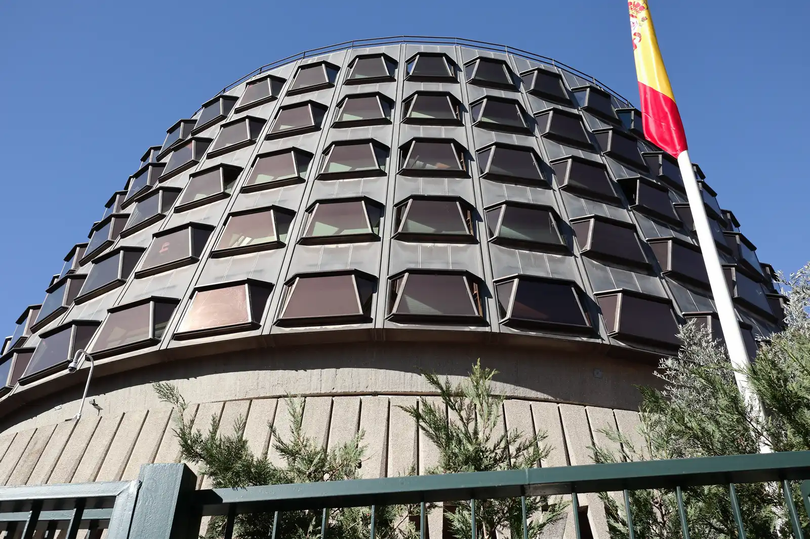 Fachada circular del Tribunal Constitucional en Madrid, de hormigón y ventanales oscuros inclinados, con la bandera de España en un mástil a la derecha y una verja verde en primer plano.