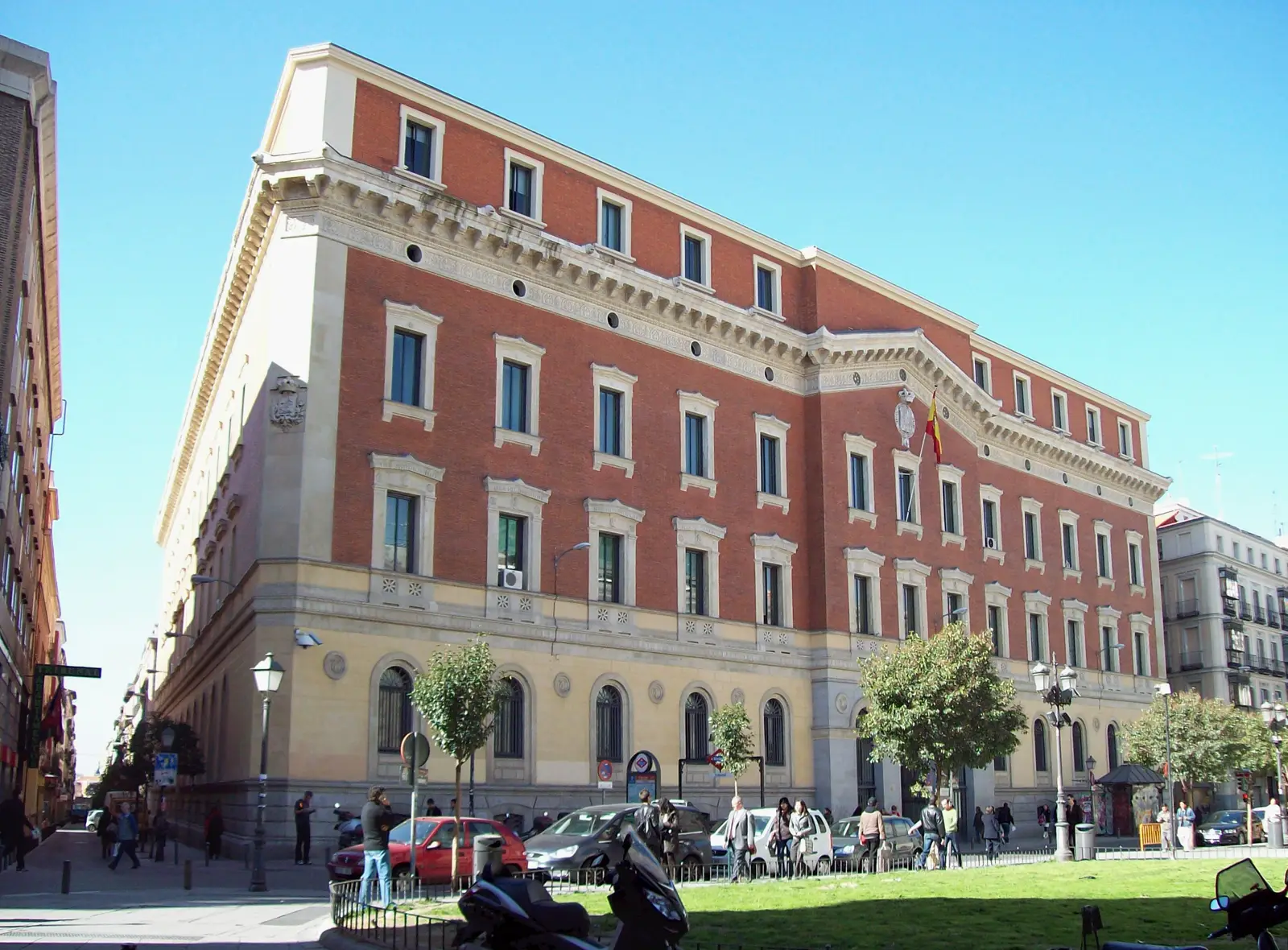 Edificio de ladrillo rojo y piedra clara de cuatro plantas visto desde una esquina, con el escudo de España y una bandera española en la fachada, coches aparcados y peatones en la acera bajo un cielo azul.
