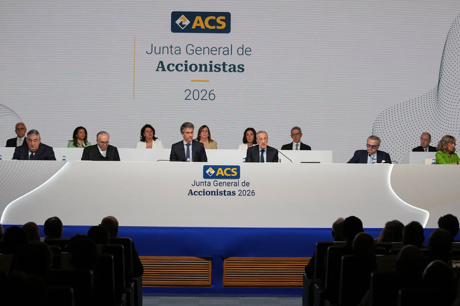Mesa presidencial de la junta general de accionistas de ACS 2026: una docena de consejeros sentados tras un estrado blanco con el logotipo de la compañía y, al fondo, una gran pantalla con el rótulo del acto.