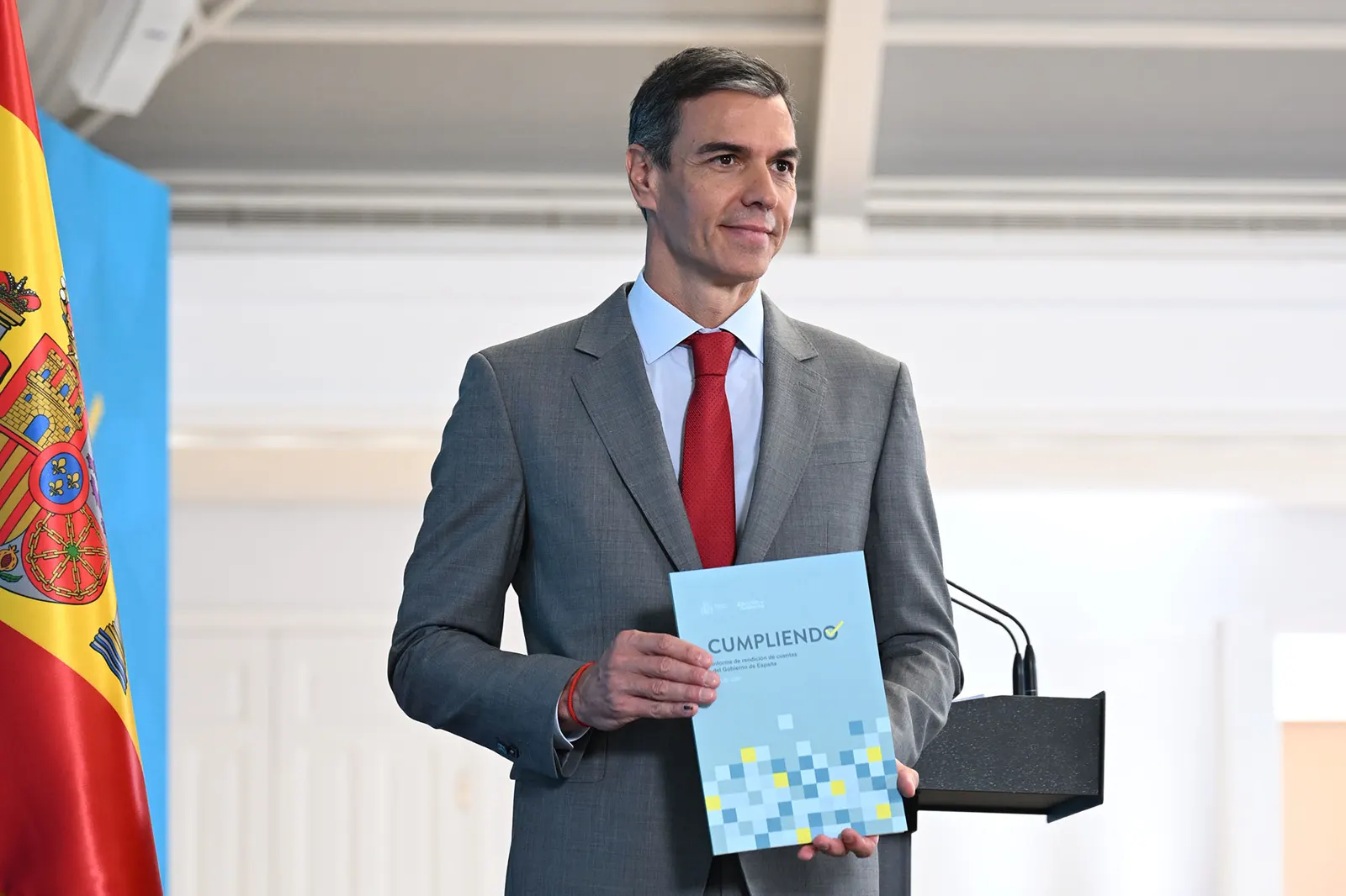 El presidente del Gobierno, Pedro Sánchez, de pie ante un atril con micrófonos, sostiene con las dos manos un documento azul titulado «Cumpliendo», con la bandera de España a su izquierda.