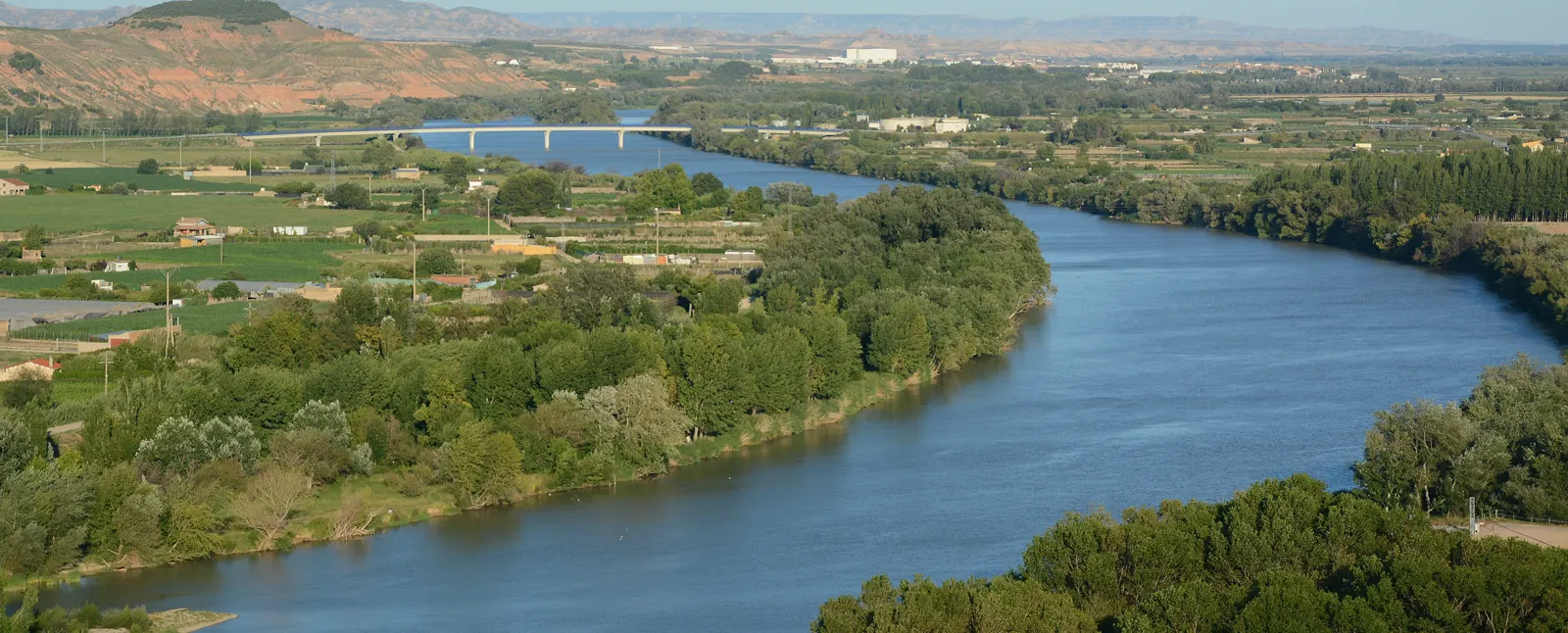 El río Ebro, ancho y de aguas tranquilas, a su paso por Tudela (Navarra), entre sotos de ribera y campos de cultivo, con un puente al fondo bajo un cielo despejado.