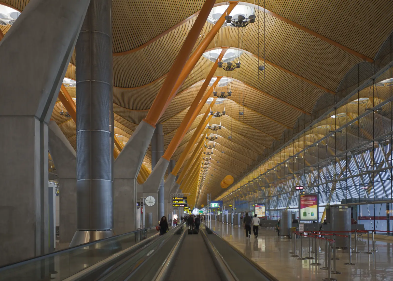 Interior de una terminal de aeropuerto con cubierta ondulada de bambú sostenida por pilares de hormigón y vigas anaranjadas; abajo, pasillos rodantes y algunos pasajeros caminando con maletas.
