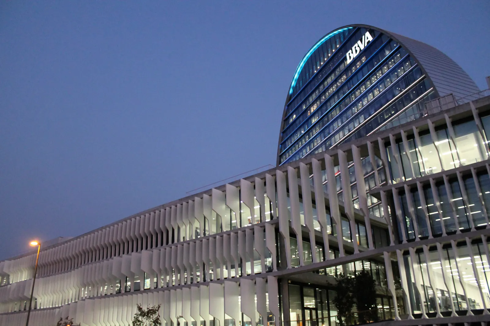 Edificio curvo acristalado con el logotipo iluminado de BBVA sobre una fachada blanca de lamas verticales, al anochecer.
