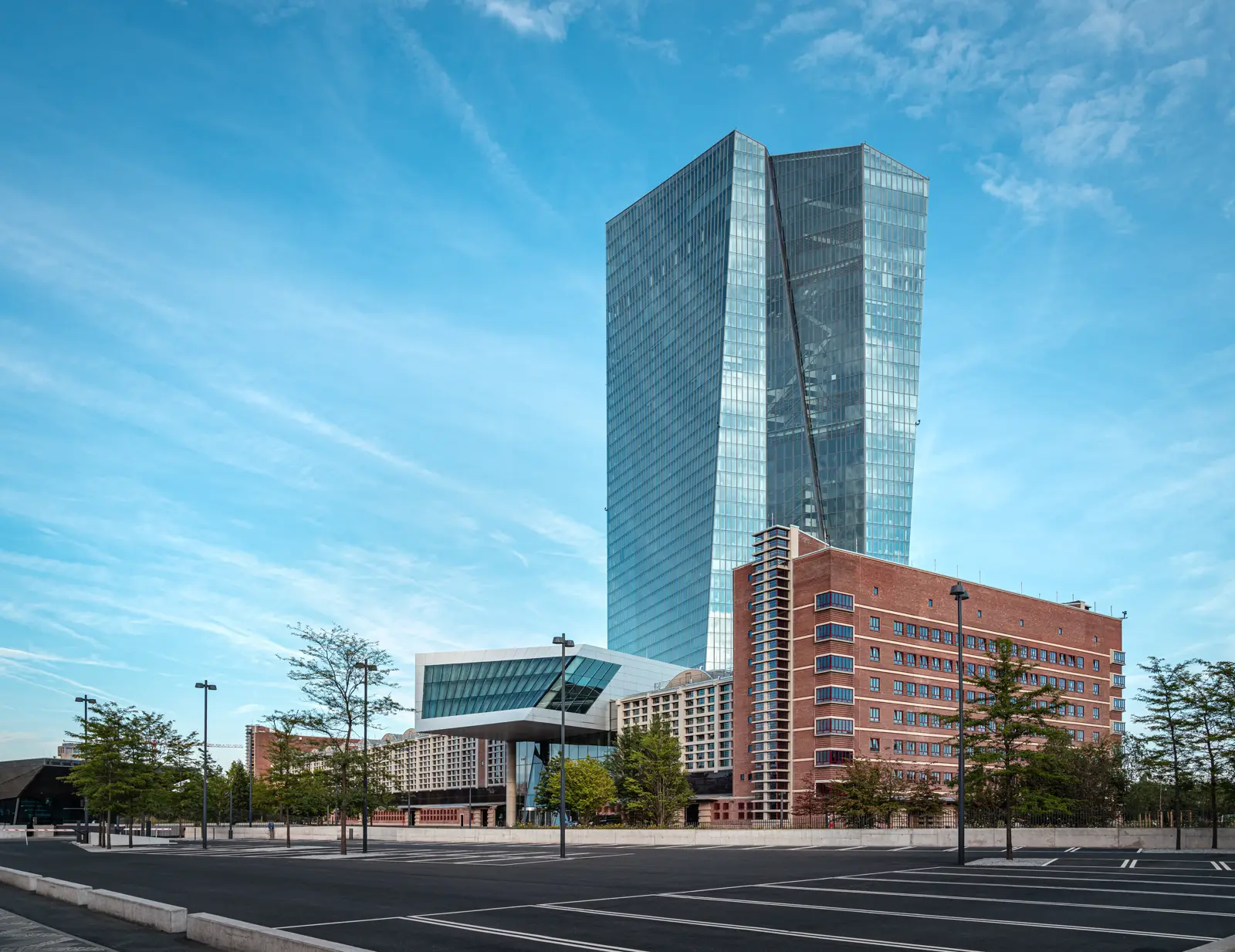 La torre acristalada del Banco Central Europeo en Fráncfort, con edificios de ladrillo rojo a sus pies y un aparcamiento vacío en primer plano, bajo un cielo azul con nubes altas