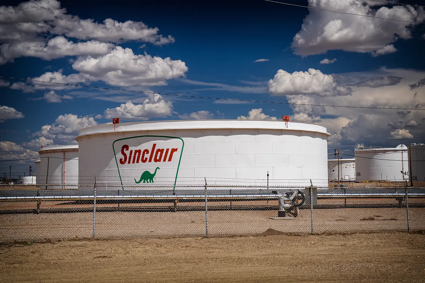 Grandes depósitos cilíndricos blancos de almacenamiento de crudo en una refinería, tras una valla metálica y bajo un cielo azul con nubes; el más cercano lleva pintado el logotipo de Sinclair.