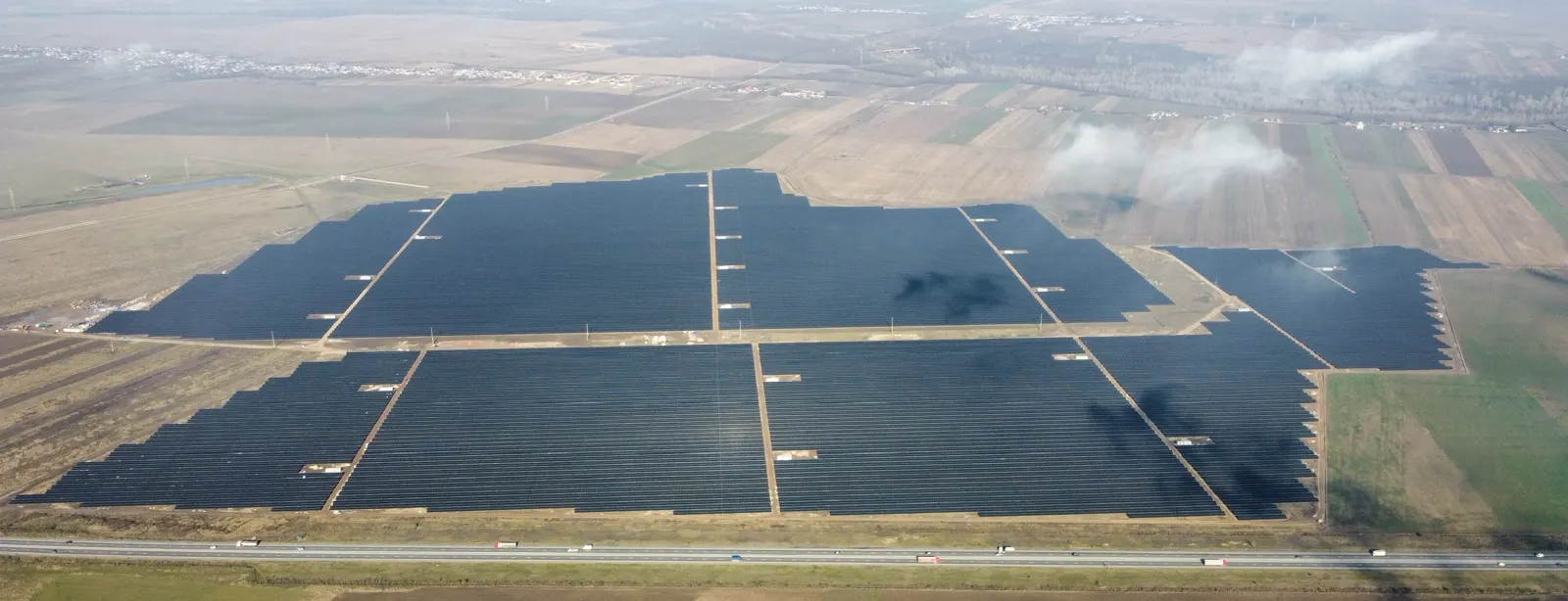 Vista aérea de un extenso parque solar fotovoltaico con filas de paneles oscuros sobre campos agrícolas de Rumanía, junto a una autopista con camiones circulando.