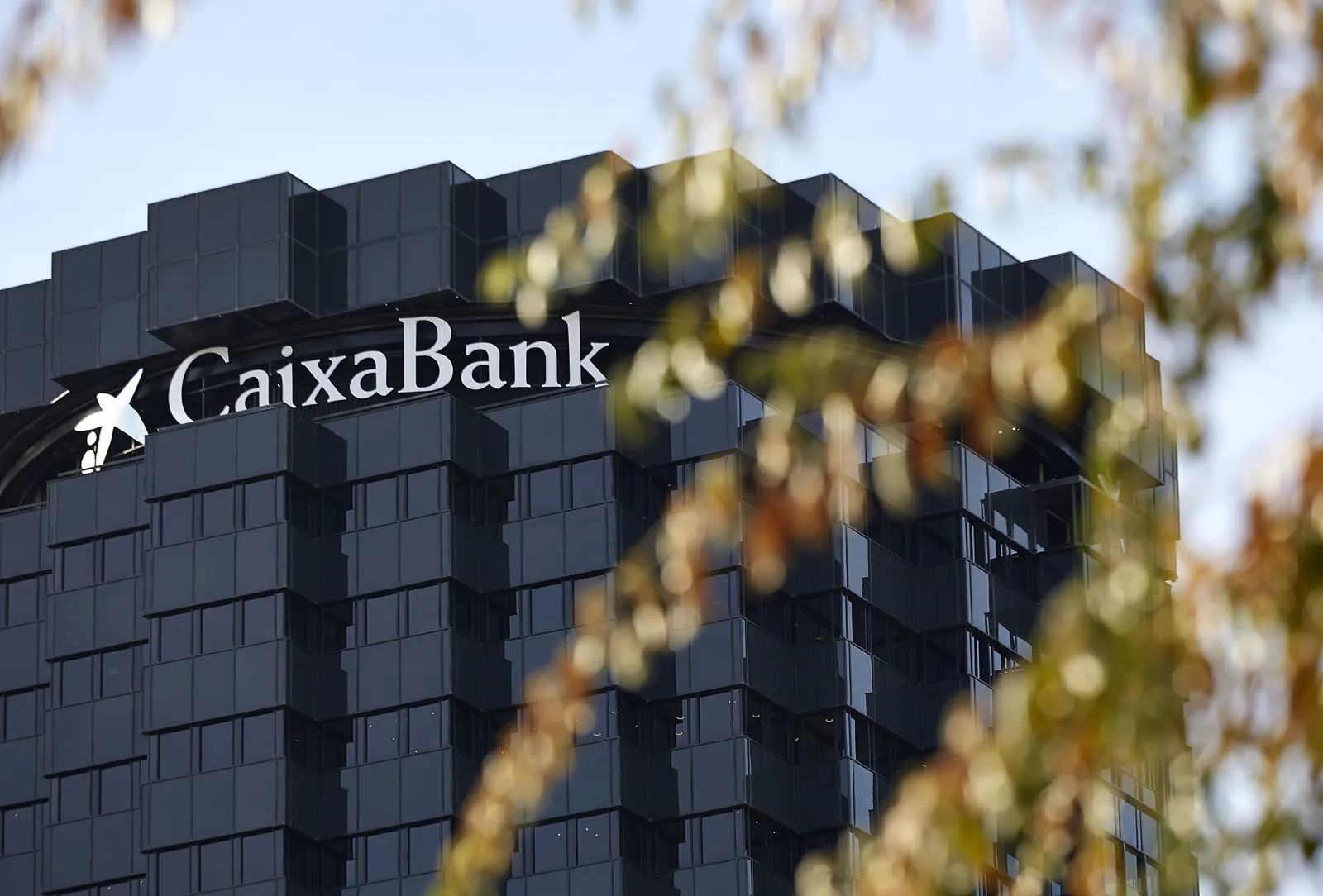 Parte superior de una torre de oficinas de fachada acristalada oscura con el rótulo y la estrella del logotipo de CaixaBank, vista entre las ramas desenfocadas de un árbol.