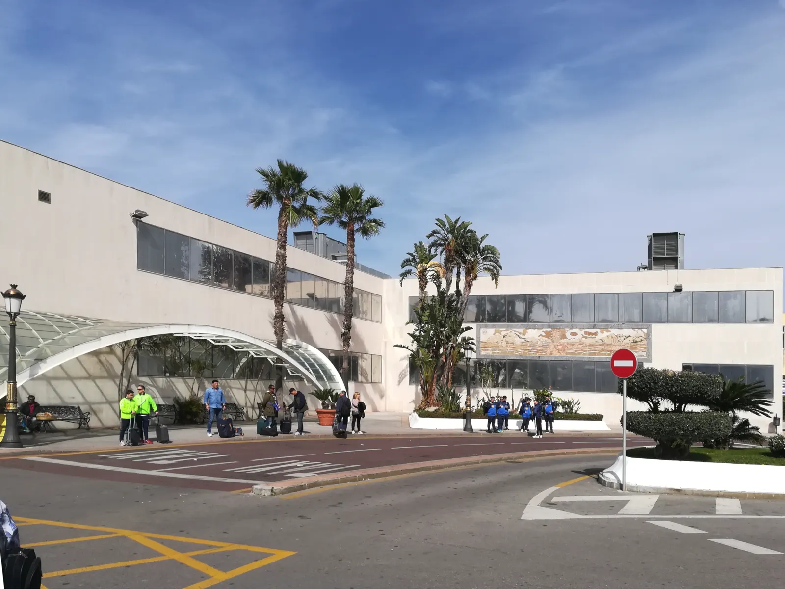 Fachada de la Estación Marítima de Ceuta, un edificio blanco de dos plantas con una marquesina acristalada sobre la entrada y palmeras delante; en la acera, varios viajeros con maletas esperan junto a la calzada