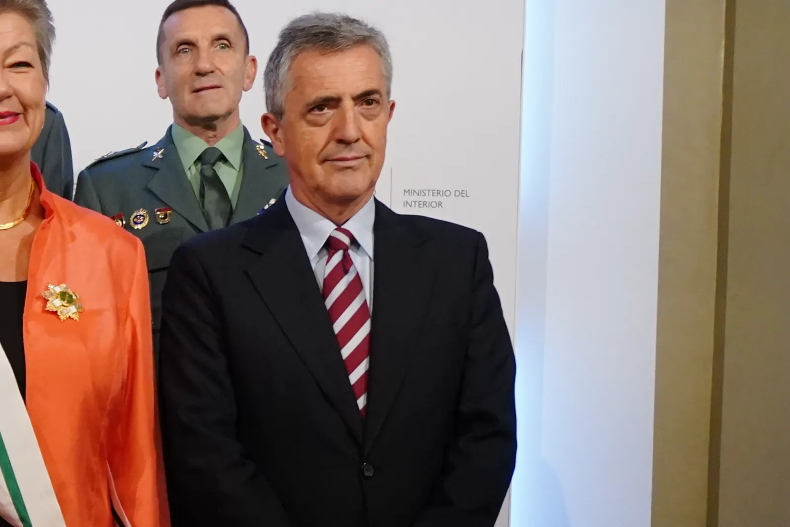 Leonardo Marcos, con traje oscuro y corbata de rayas granates y blancas, posa ante un panel con el rótulo «Ministerio del Interior»; detrás, un mando de la Guardia Civil de uniforme y, a la izquierda, una mujer con chaqueta naranja y banda de condecoración.