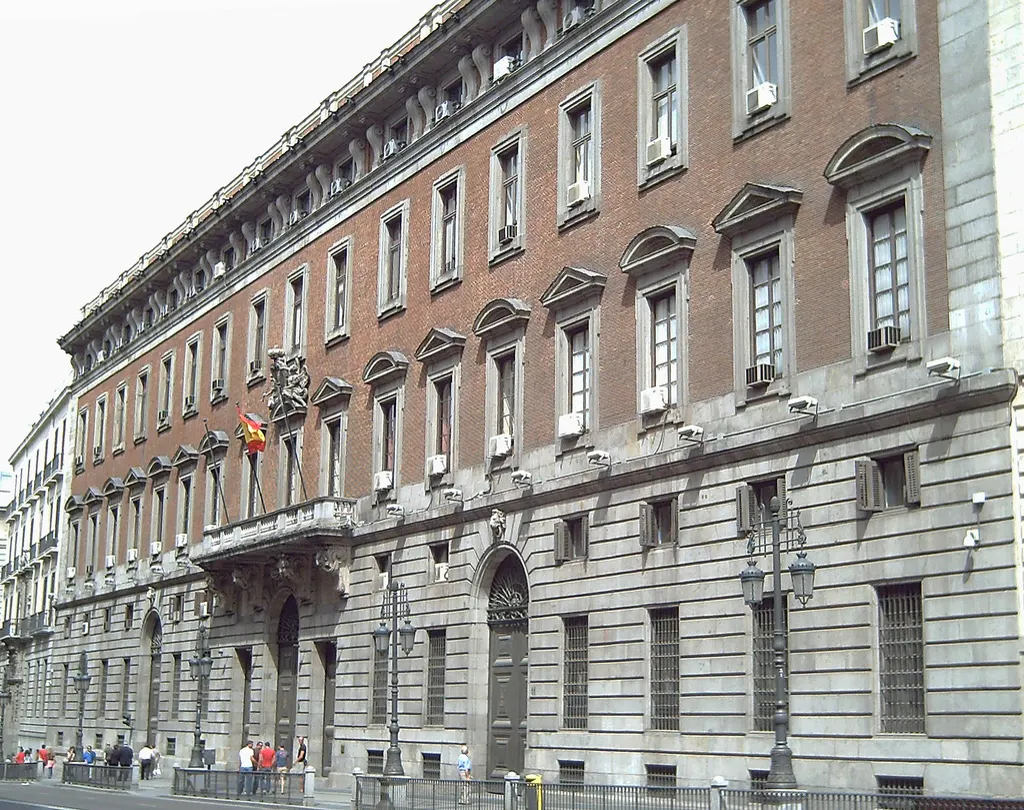 Fachada de la Real Casa de la Aduana, edificio de ladrillo y piedra en la calle de Alcalá de Madrid, con la bandera de España sobre el balcón principal y viandantes en la acera.