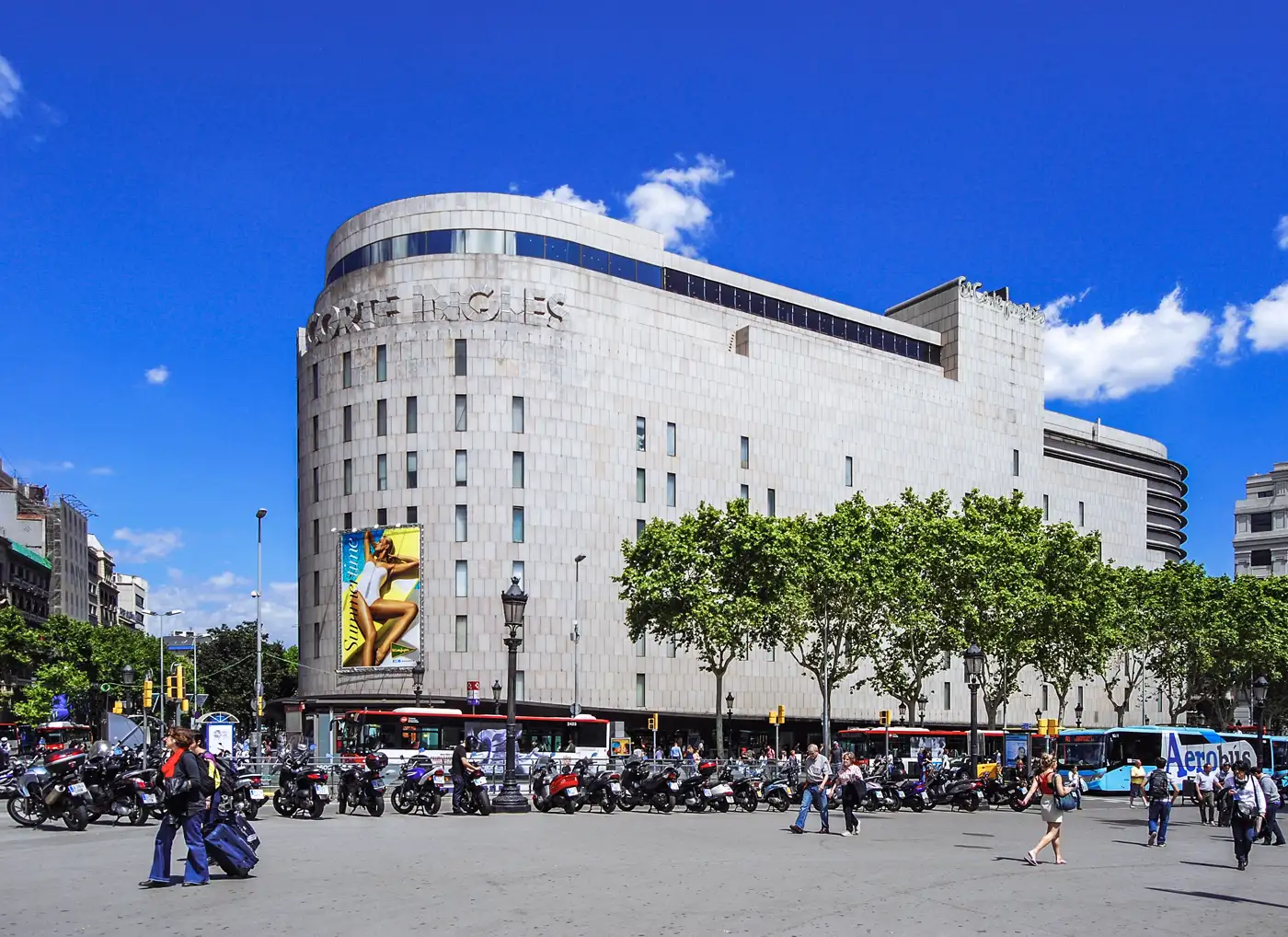 Edificio de El Corte Inglés en la plaza de Cataluña de Barcelona, de piedra clara y esquina redondeada, con árboles, motos aparcadas y peatones cruzando la plaza en primer plano bajo un cielo azul.