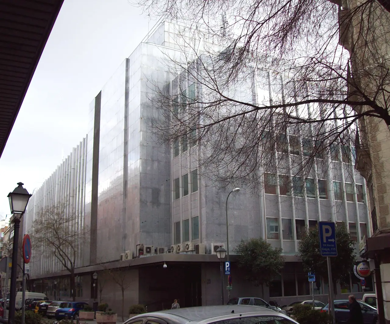 Edificio acristalado de varias plantas visto desde la acera de enfrente, con fachada de vidrio y aletas verticales blancas, árboles sin hojas y coches aparcados en una calle de Madrid.