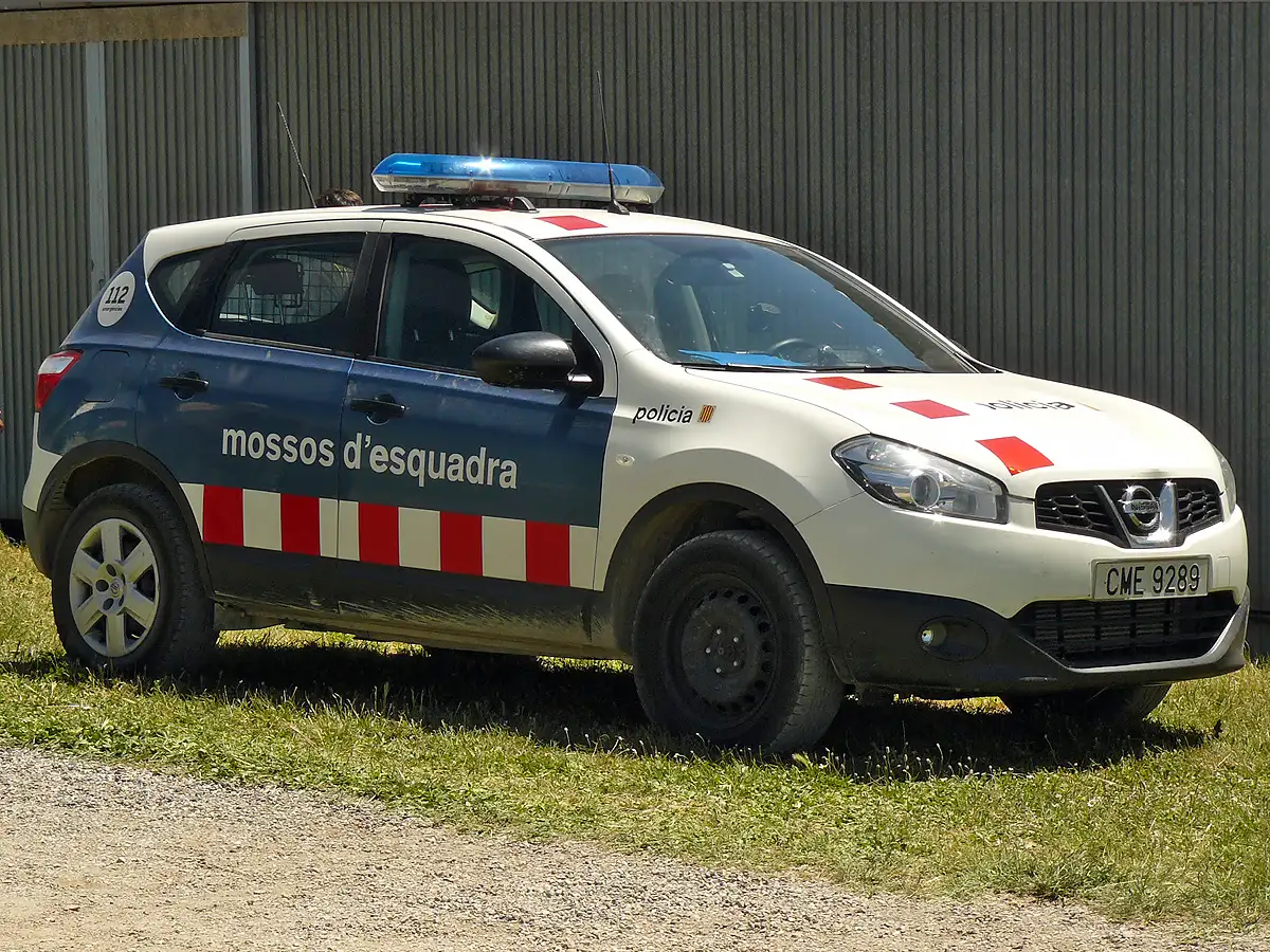 Coche patrulla blanco y azul con el rótulo «mossos d'esquadra» en el lateral, aparcado sobre hierba junto a una nave de chapa