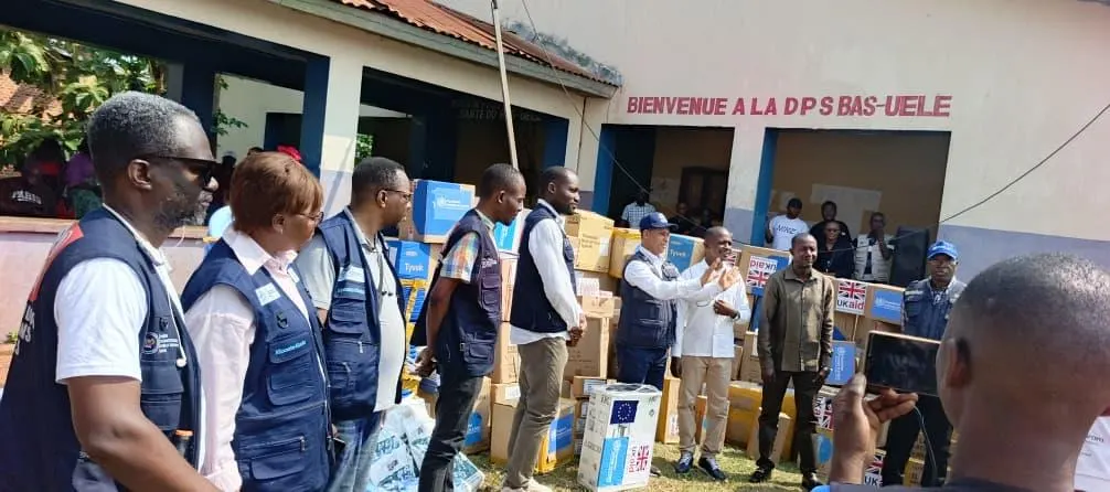 Una fila de sanitarios y responsables con chalecos de identificación escucha a un hombre que habla junto a decenas de cajas de material médico apiladas ante un edificio con el rótulo «Bienvenue a la DPS Bas-Uélé»