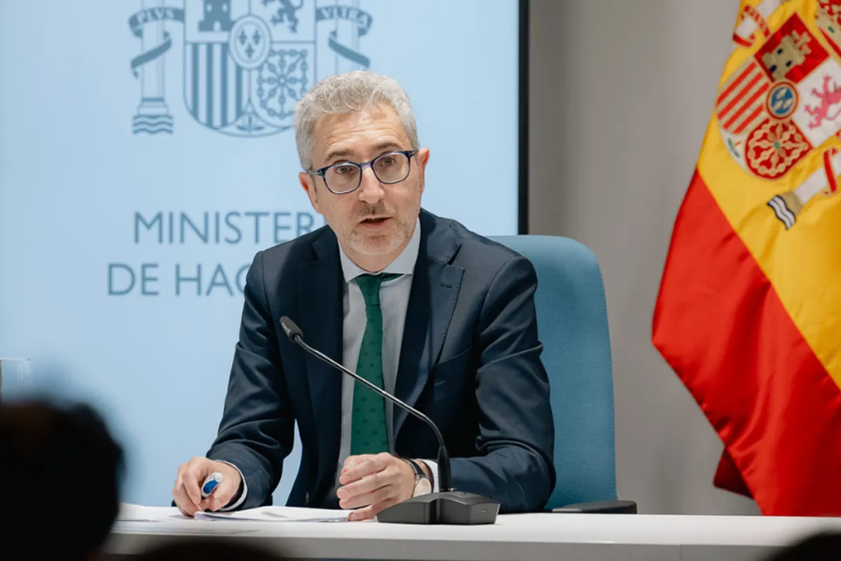 El ministro de Hacienda, Arcadi España, sentado ante un micrófono durante una rueda de prensa, con el logotipo del Ministerio de Hacienda al fondo y una bandera de España a su derecha.