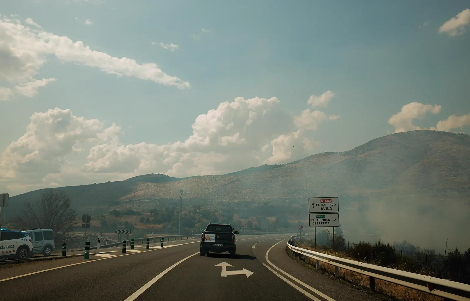 Humo del incendio cubriendo la carretera N-403, en la provincia de Ávila, con señales hacia El Barraco y Ávila y la salida hacia El Tiemblo y Cebreros; un coche de la Policía Local cierra un acceso y una pickup circula entre el humo.