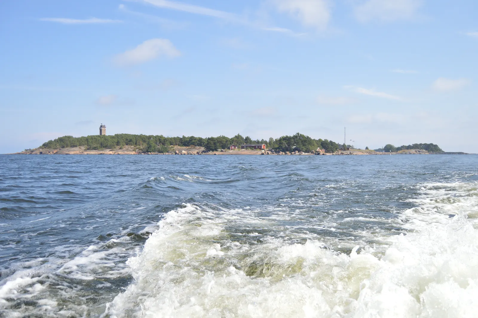 Isla baja y boscosa vista desde el mar, con una torre de faro de piedra a la izquierda, casas de madera rojas junto a una orilla rocosa y la estela de espuma de una embarcación en primer plano.