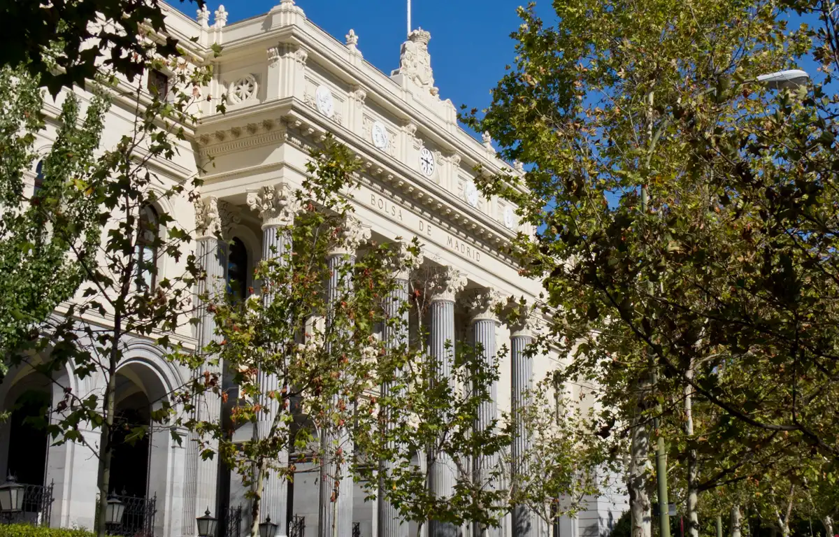 Fachada neoclásica del Palacio de la Bolsa de Madrid, de piedra clara con columnas y bandera de España en lo alto, parcialmente cubierta por árboles bajo un cielo azul.