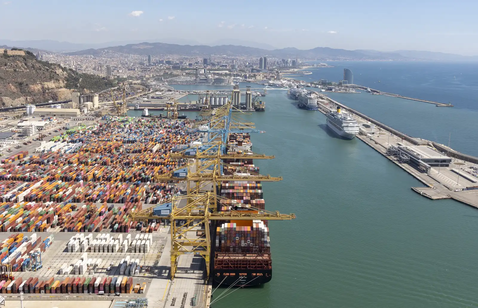Vista aérea de la terminal de contenedores del puerto de Barcelona: miles de contenedores de colores apilados, grúas pórtico amarillas cargando un portacontenedores y, al fondo, la ciudad y el muelle de cruceros.