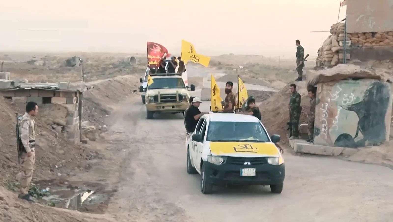 Convoy de camionetas con banderas amarillas y milicianos armados avanza por una pista de tierra junto a un puesto fortificado con sacos terreros.