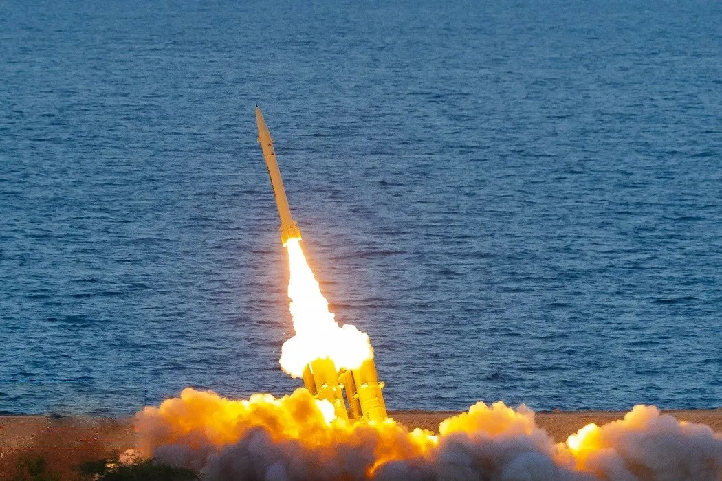 Un misil se eleva sobre una columna de fuego y humo desde un lanzador situado en la costa, con el mar de fondo.