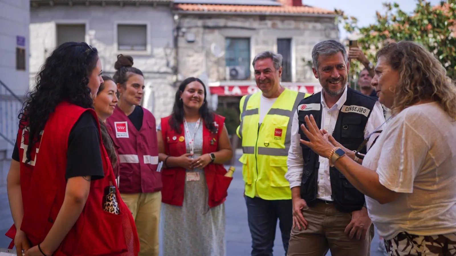 Un hombre con chaleco caqui rotulado «Consejero» conversa sonriente con una vecina y con cinco voluntarios con chalecos rojos y amarillo de la Comunidad de Madrid, en la plaza de un pueblo con casas de piedra al fondo.
