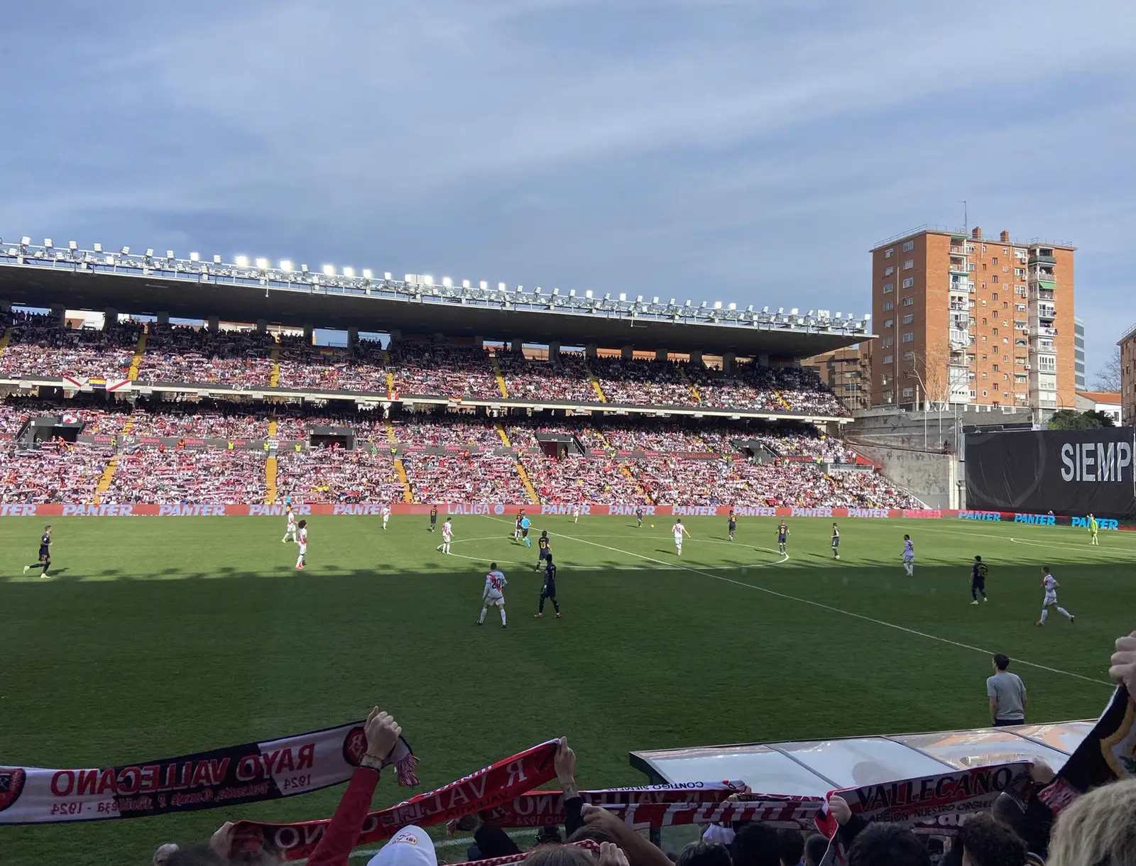 Gradas llenas y césped del Estadio de Vallecas durante un partido del Rayo Vallecano, con aficionados alzando bufandas del club en primer plano.