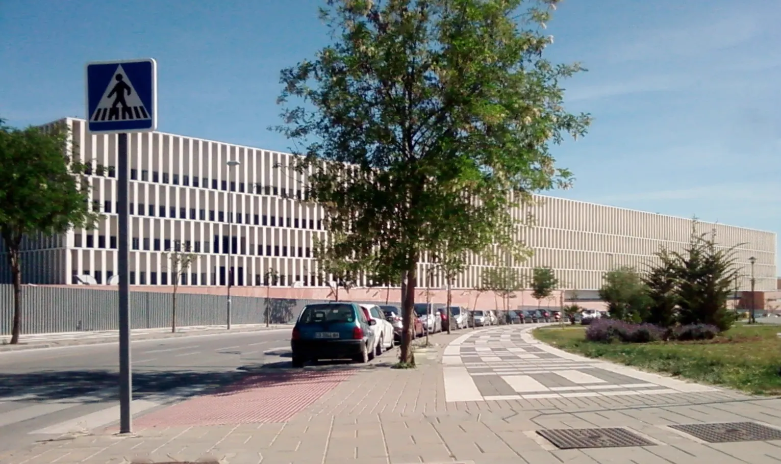 Edificio judicial alargado de fachada blanca con lamas verticales, visto desde una calle con coches aparcados, árboles y una señal de paso de peatones en primer plano.
