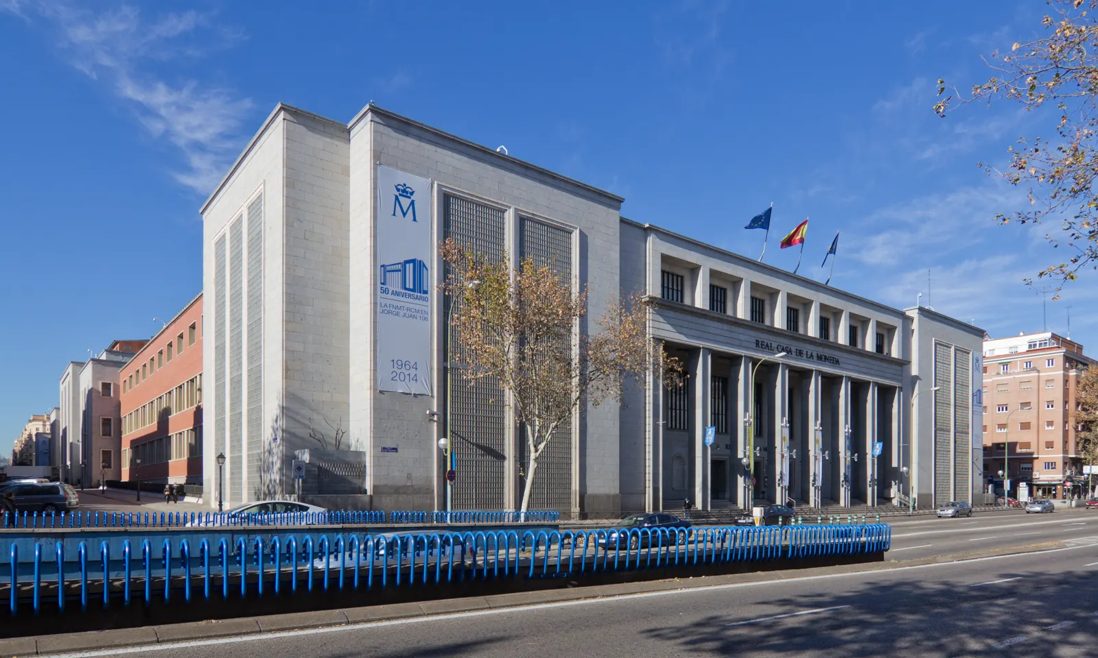 Fachada de piedra clara de la Real Casa de la Moneda en Madrid, con la leyenda REAL CASA DE LA MONEDA sobre la columnata y banderas de España y la Unión Europea