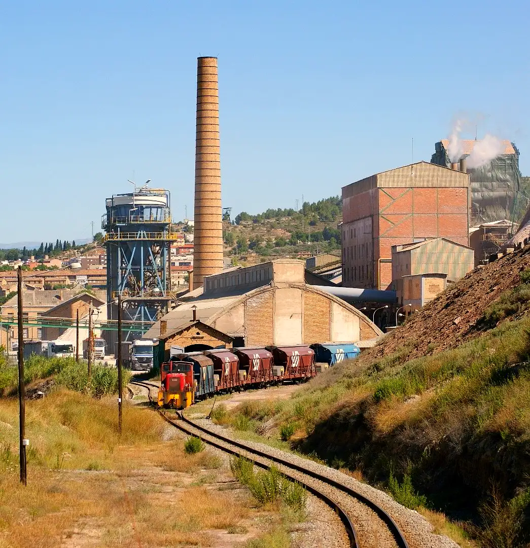 Instalaciones mineras de la potasa en Súria: torre de extracción metálica, chimenea de ladrillo, naves industriales y una locomotora con tolvas de potasa en primer término.