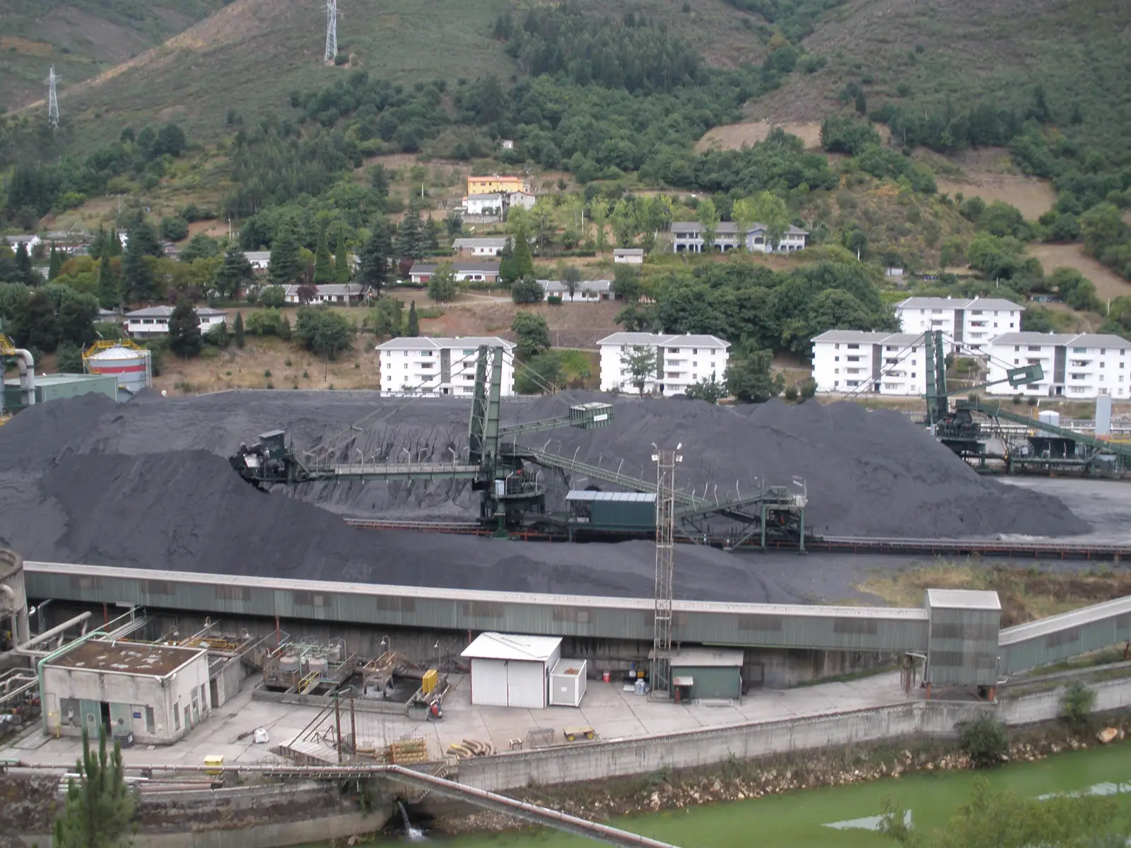 Parque de carbones de la central térmica del Narcea: montañas de carbón, cintas transportadoras y apiladoras verdes, con bloques de viviendas y ladera boscosa al fondo