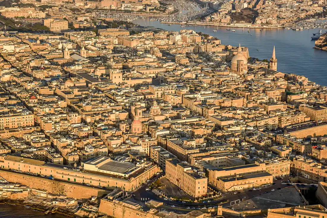 Vista aérea de La Valeta al atardecer: manzanas densas de edificios de piedra caliza con azoteas planas, dos cúpulas de iglesia y un campanario, rodeadas de murallas y con el puerto y el mar al fondo.