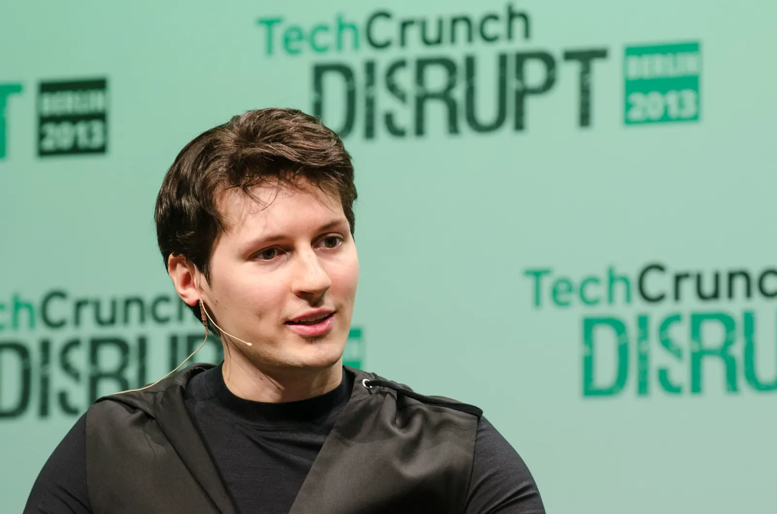 Hombre joven de pelo oscuro y camiseta negra, con micrófono de diadema, hablando sentado ante un panel verde con el rótulo «TechCrunch Disrupt Berlin 2013».