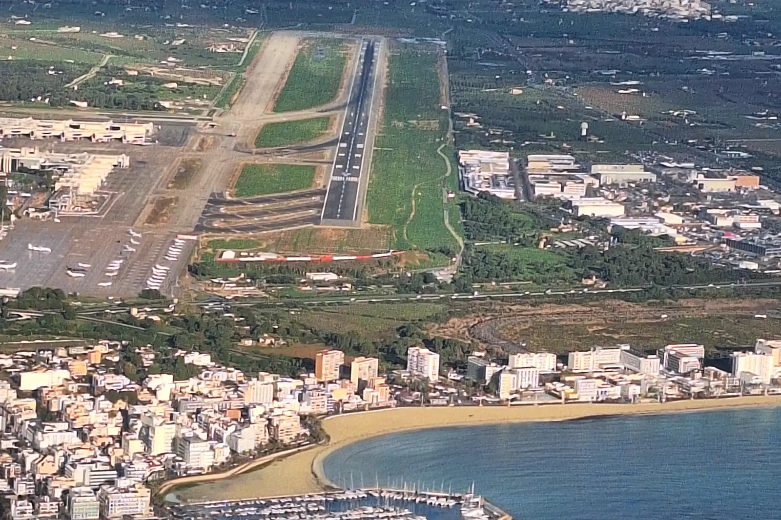 Vista aérea del aeródromo de Son Sant Joan, en Palma: una pista de aterrizaje en diagonal, una amplia plataforma con decenas de aviones estacionados junto a las terminales y, al fondo, los bloques de Can Pastilla, la playa y el puerto deportivo sobre el mar.