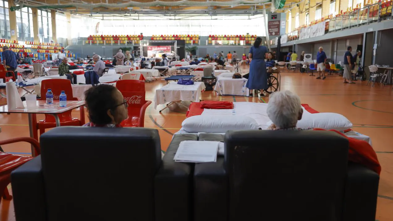 Interior de un polideportivo con camas plegables, mantas y almohadas; en primer plano, dos personas mayores sentadas de espaldas y, al fondo, vecinos y personal de atención entre las gradas.