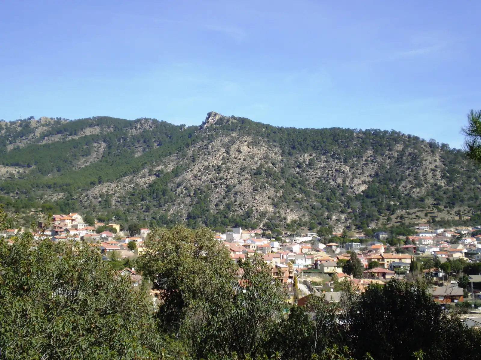 Vista general de Valdemaqueda (Madrid): caserío de tejados rojizos rodeado de arbolado al pie de una ladera de granito cubierta de pinar, bajo un cielo despejado.