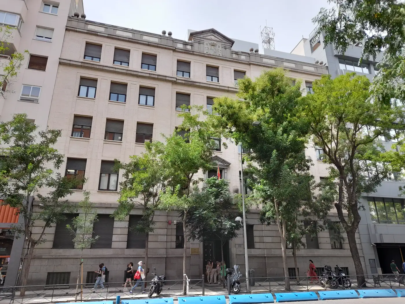 Fachada de piedra clara de un edificio de cinco alturas con un escudo esculpido en el frontón superior; en la planta baja, una puerta con bandera de España, y delante una acera con árboles, viandantes y motos aparcadas.