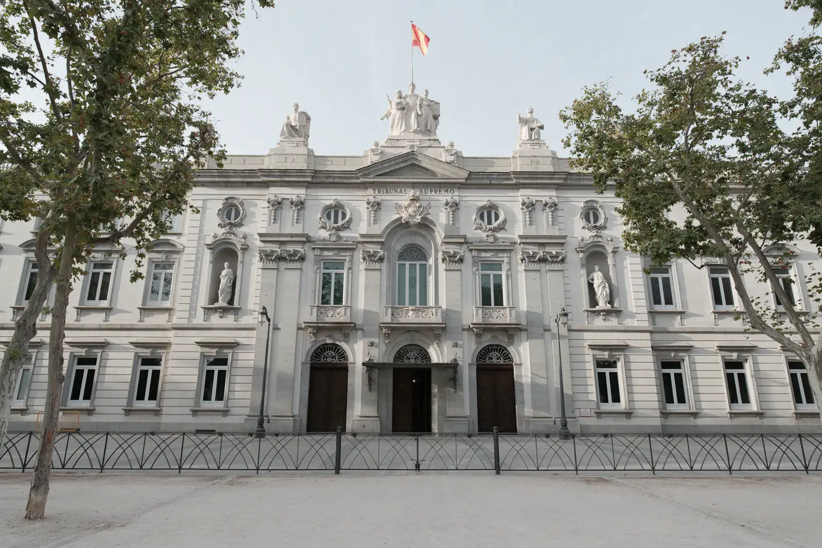Fachada neoclásica de piedra gris del Tribunal Supremo en Madrid, con la inscripción «Tribunal Supremo» bajo el frontón, grupos escultóricos en el remate, estatuas de juristas en hornacinas, tres portones de madera, una verja negra de arcos entrelazados delante y la bandera de España en lo alto, entre las copas de varios árboles.