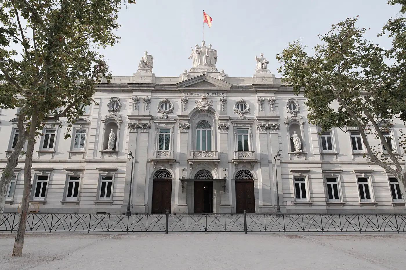Fachada neoclásica de piedra clara del Tribunal Supremo en Madrid, con la inscripción «Tribunal Supremo» bajo el frontón, estatuas de juristas en hornacinas, bandera de España ondeando en el tejado y una verja negra delante, enmarcada por dos plátanos de sombra.