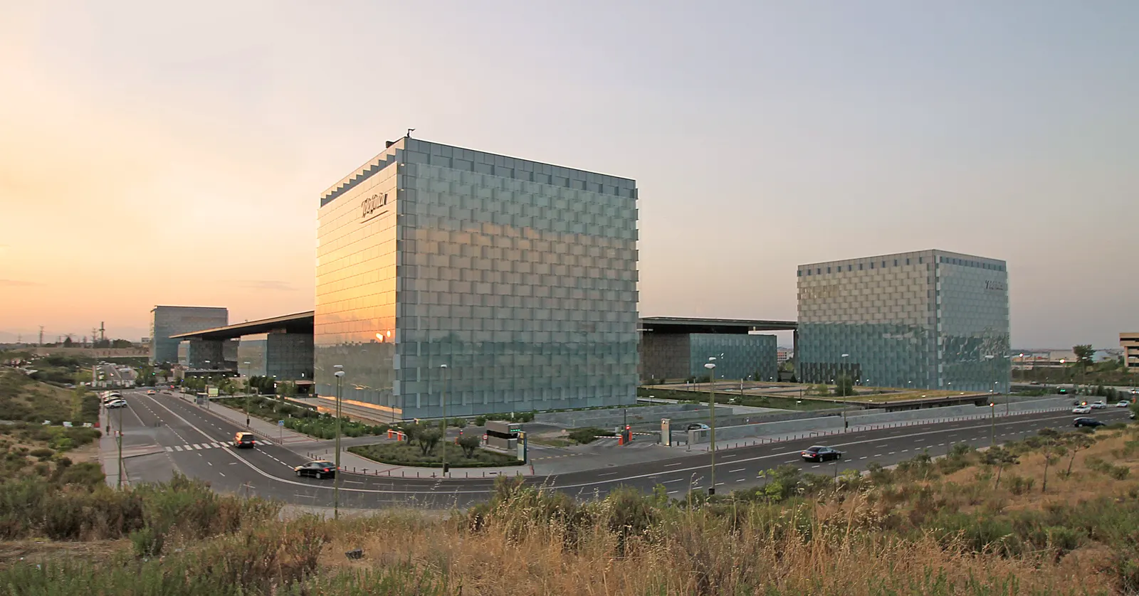 Vista al atardecer de dos grandes edificios acristalados de planta cuadrada rodeados de avenidas con tráfico, con el logotipo de Telefónica en lo alto de la fachada.