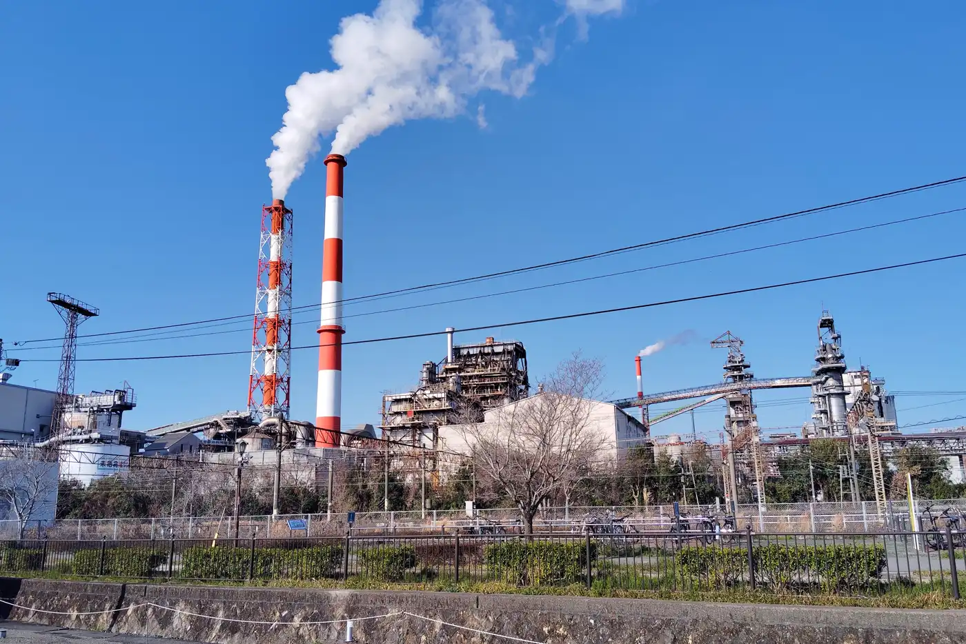 Vista general de la fábrica de Nippon Paper en Yatsushiro bajo un cielo azul: dos chimeneas altas pintadas a franjas rojas y blancas expulsan columnas de vapor sobre las naves industriales, con árboles y una valla en primer plano.