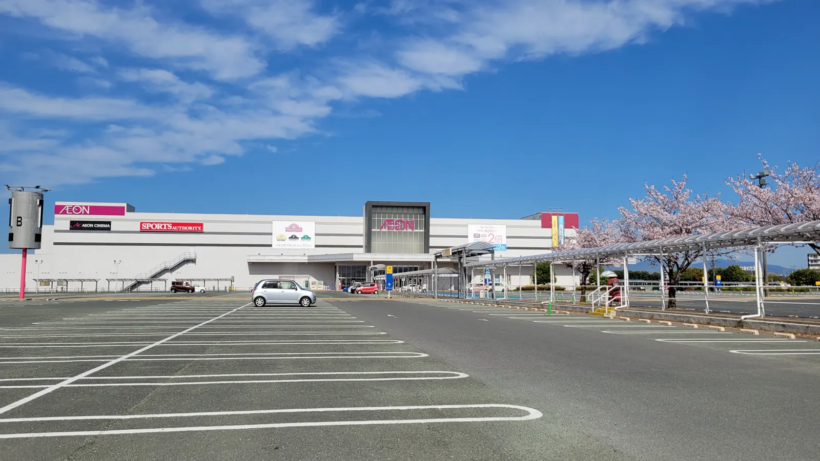 Fachada del centro comercial AEON Mall Kumamoto vista desde su aparcamiento vacío, un edificio blanco alargado de dos plantas con el rótulo rosa de AEON, bajo un cielo azul con nubes y con un cerezo en flor a la derecha.