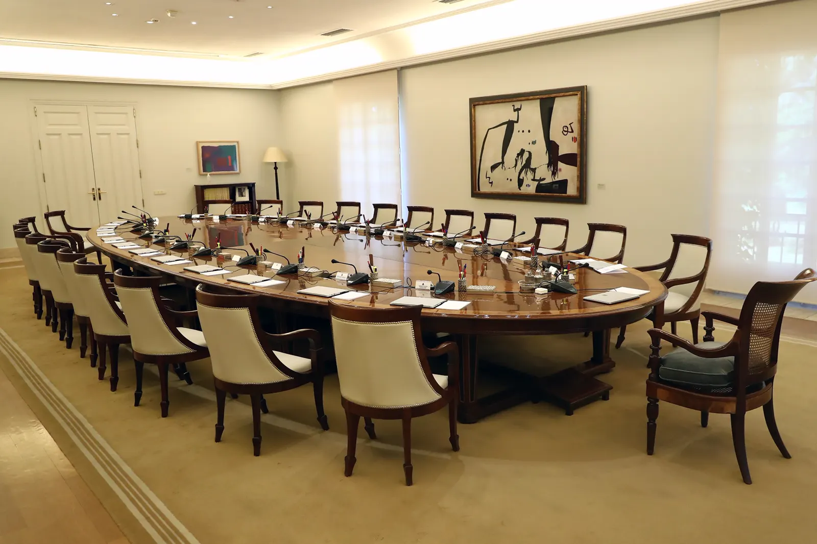 Sala vacía con una gran mesa ovalada de madera rodeada de sillas tapizadas en blanco, con micrófonos, carpetas y bloques de notas sobre el tablero, y dos cuadros abstractos en las paredes.
