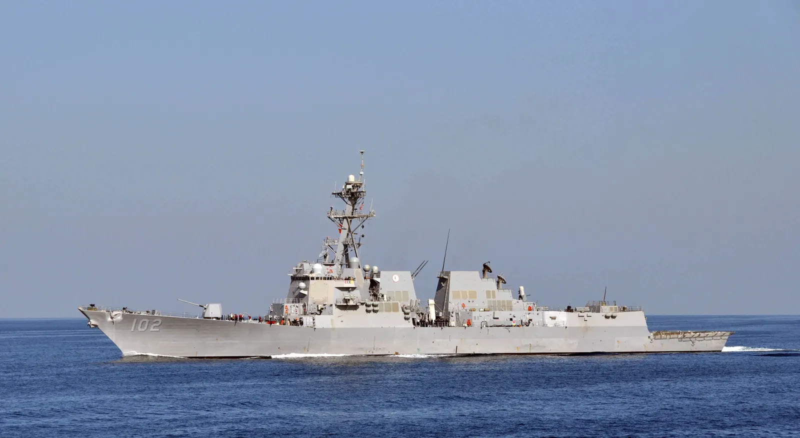 Un destructor de guerra gris con el número 102 en la proa navega solo en mar abierto bajo un cielo despejado; en cubierta se distinguen varios marineros con chalecos de color.