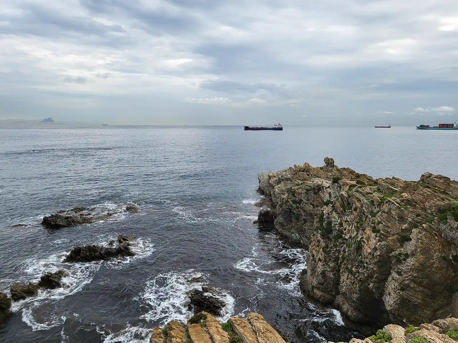 Costa rocosa del norte de Ceuta: rompientes de espuma sobre las rocas en primer término, el mar en calma y, en el horizonte, varios cargueros fondeados y el perfil de la costa al otro lado del Estrecho bajo un cielo nublado