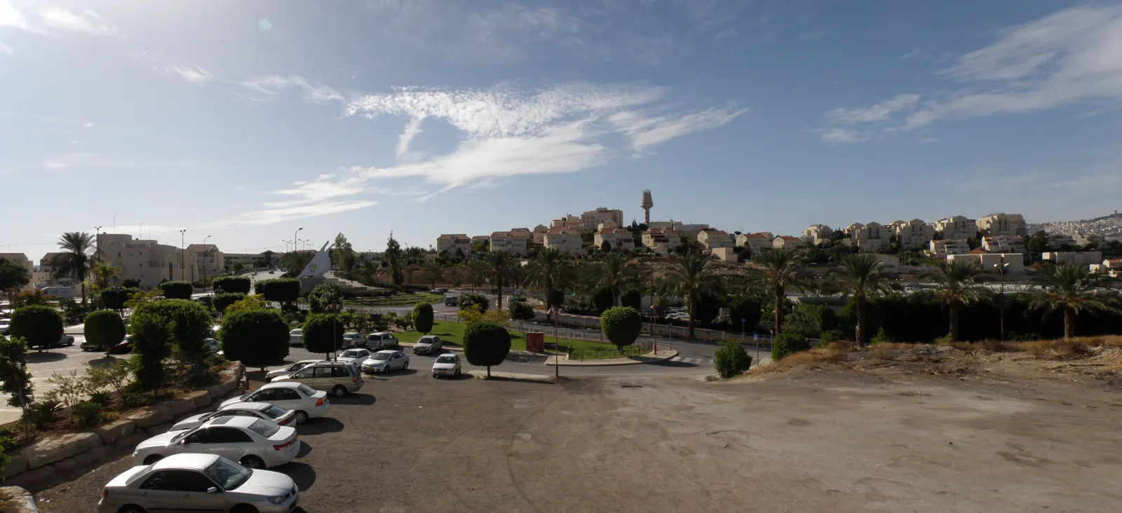 Vista de las viviendas del asentamiento israelí de Maale Adumim, en Cisjordania, sobre una ladera desértica, con palmeras y un aparcamiento en primer plano.