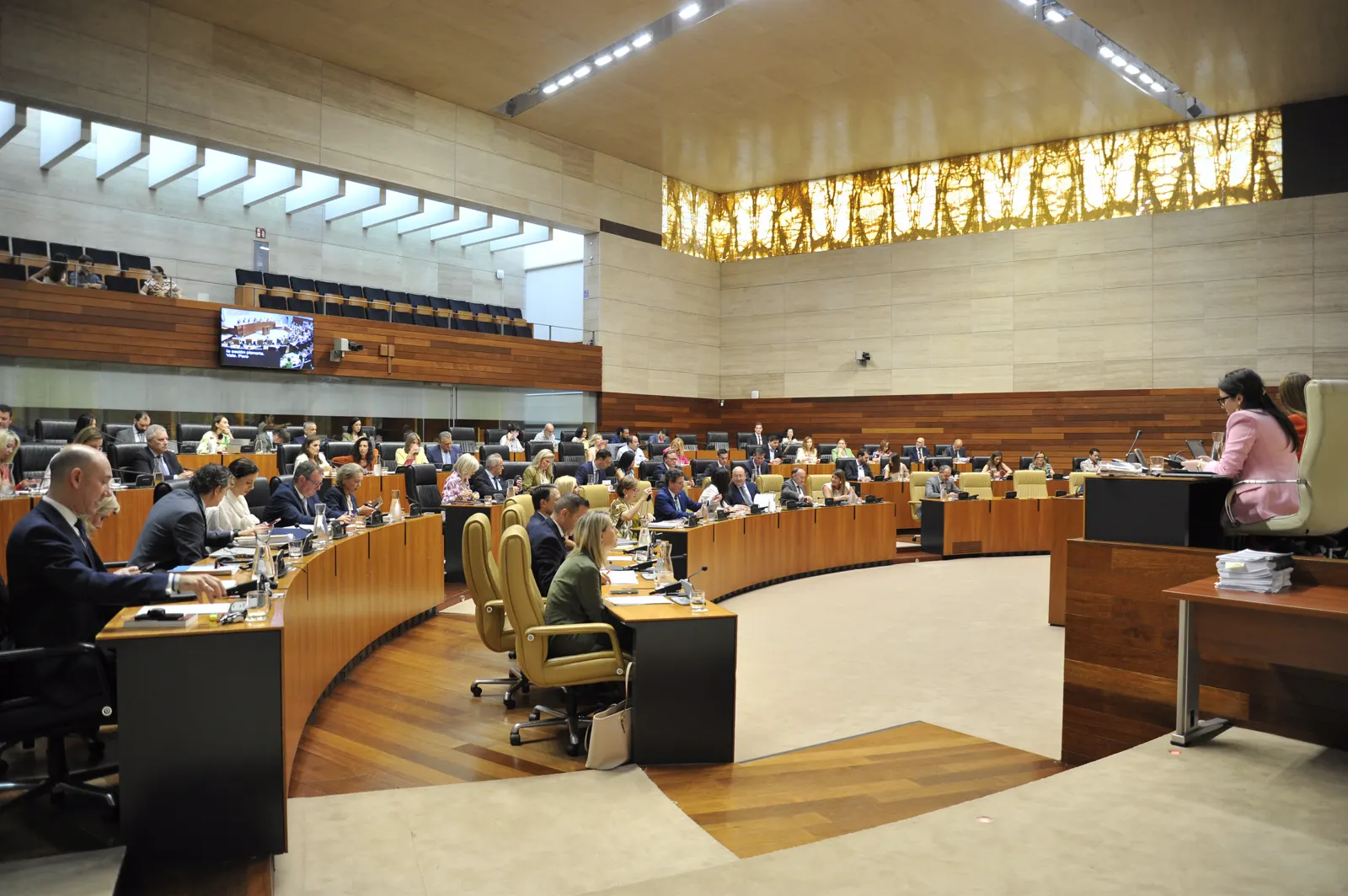Vista general del hemiciclo de la Asamblea de Extremadura con los escaños ocupados por diputados durante una sesión plenaria; a la derecha, una diputada interviene desde la tribuna de oradores.